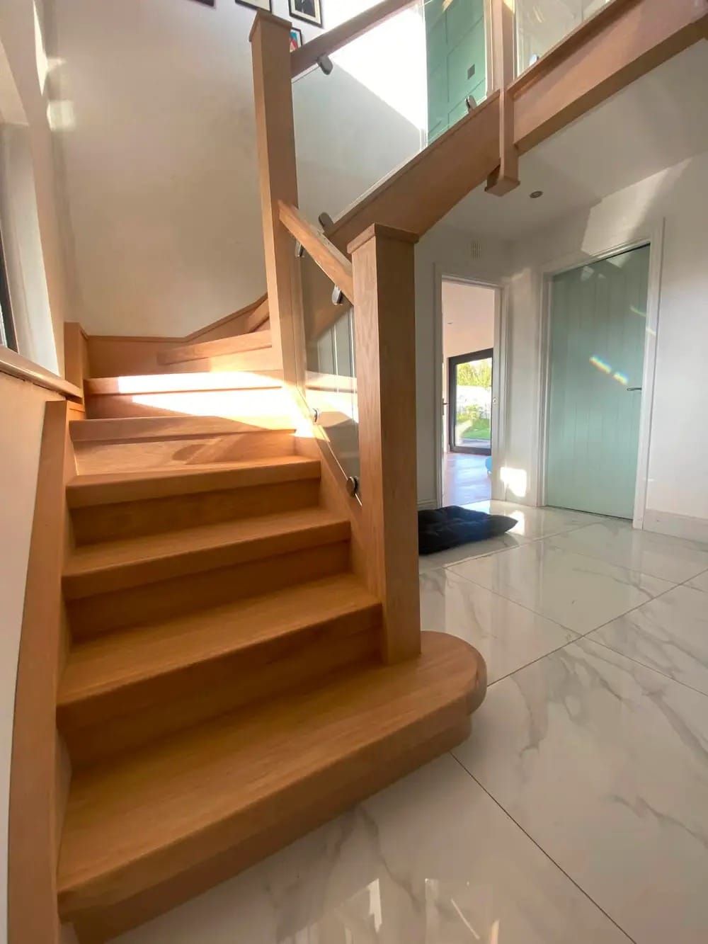 Solid oak winder staircase with glass balustrade panels in a sunlit marble-tiled hallway